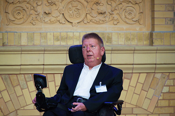 Ein Mann im Anzug, der im Rollstuhl sitzt, spricht in ein Mikrofon. Er trägt einen schwarzen Anzug und ein weißes Hemd. Er hat kurze, blonde Haare.