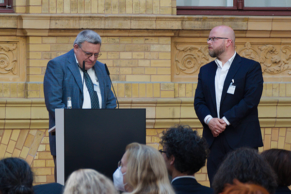 Zwei Männer stehen auf der Bühne. Einer der beiden steht hinterm Rednerpult. Er hat kurze graue Haare und trägt eine Brille. Der andere Mann hat eine Glatze und trägt einen Vollbart und ebenfalls eine Brille. Er sieht zu dem Mann am Rednerpult hinüber.