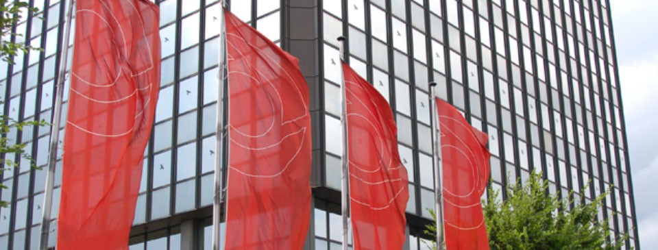 Das Bild zeigt das Gebäude der Knappschaft-Bahn-See in Bochum. Davor wehen Fahnen mit dem Logo der Knappschaft auf rotem Grund.