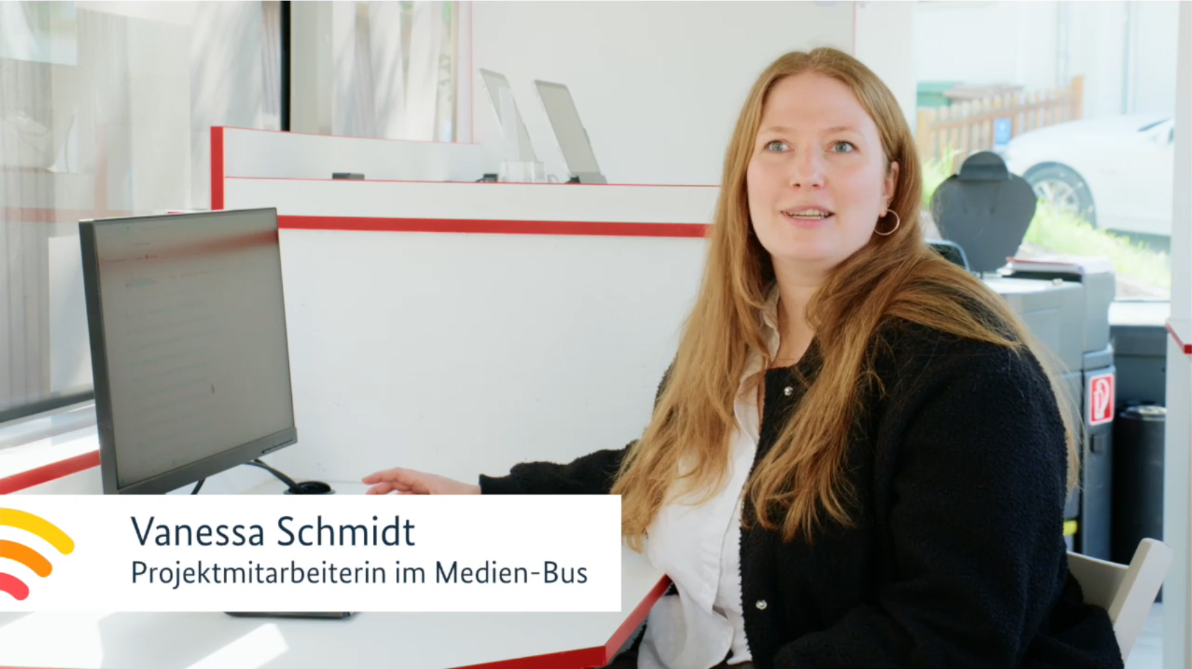 Eine Frau mit langen rötlichen Haaren spricht. Sie sitzt am Schreibtisch in einem Bus.