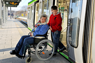 Eine Frau in einem Rollstuhl wird von einem Jungen – vielleicht ihr Sohn – aus einem Regionalzug geschoben. Beide schauen zurück auf den niveaugleichen Übergang zwischen Zug und Bahnsteigkante. Auf dem Bahnsteig ist ein Bodenleitsystem verlegt.
