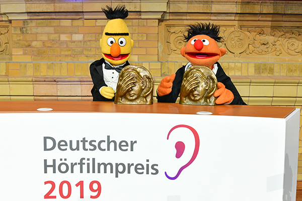 Ernie und Bert stehen hinter dem Podest des Deutschen Hörfilmpreises, vor ihnen steht jeweils der Preis: die Bronzeskulptur ADele | Foto: DBSV/Oliver Ziebe