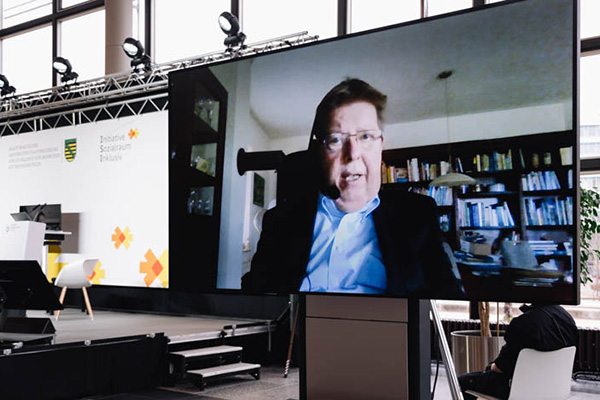 Dr. Volker Sieger auf einem Bildschirm, der neben der Bühne steht- Foto: Bundesfachstelle Barrierefreiheit/Carsten Beier