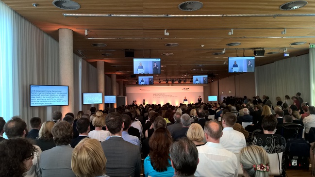 Eine Zuschauermenge sitzt vor einem Podium. Auf diesem befinden sich 4 Personen, Gebärden- und Schriftdolmetscher und eine Referentin. Im Raum verteilt sind Monitore zum Verfolgen der Veranstaltung in Gebärdensprache und Schriftmittlung.