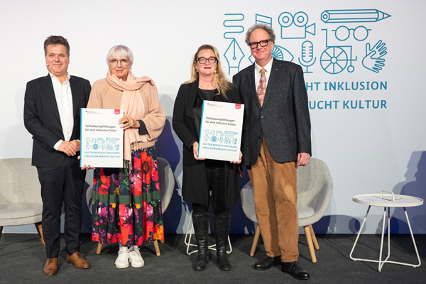 Zwei Männer und zwei Frauen stehen auf einer Bühne. Sie halten zwei große Dokumente in der Hand: "Teilhabeempfehlungen für eine inklusive Kultur".