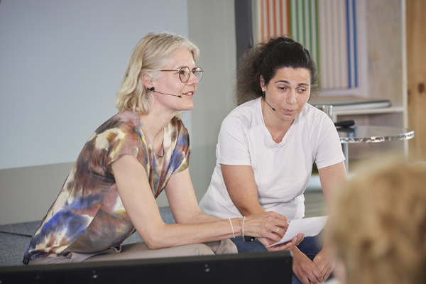 Zwei Frauen sitzen auf dem Rand einer Bühne. Beide tragen Kopfmikrofone und sprechen.