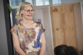 Eine Frau spricht in ein Kopfmikrofon. Sie hat schulterlange blonde Haare und trägt eine Brille.
