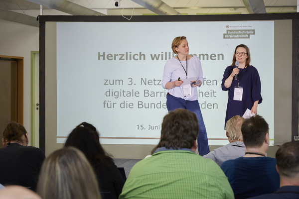 Zwei Frauen stehen auf einer Bühne vor einer Leinwand, eine der Frauen spricht in ein Mikrofon. Im Hintergrund steht: Herzlich Willkommen.
