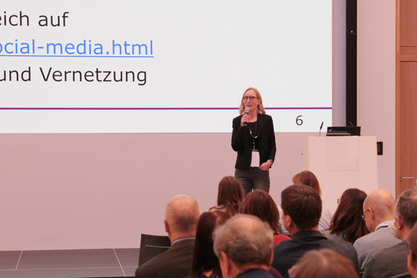 Eine Frau mit blonden Haaren und Brille steht auf einer Bühne und spricht in ein Mikrofon. Davor ist Publikum zu sehen, dahinter die Leinwand mit ihrer Präsentation.