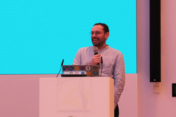 Ein Mann steht an einem Pult und spricht in ein Mikrofon. Er hat kurze dunkle Haare und trägt eine Brille und einen Bart.