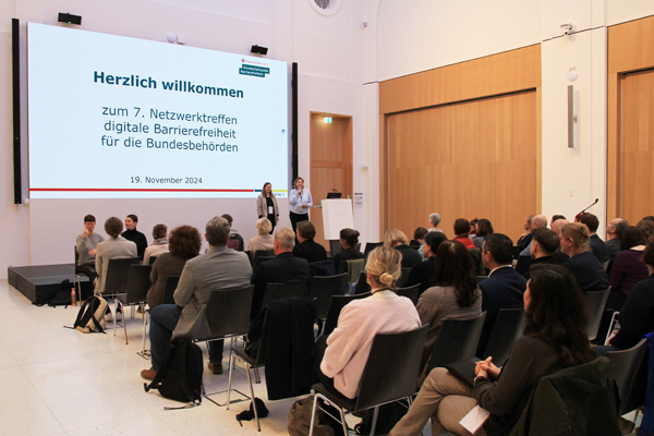 Blick in den Raum: Viele Teilnehmerinnen und Teilnehmer sitzen im Raum, vorne auf der Bühne stehen zwei Frauen. Dahinter ist ein "Herzlich willkommen" zu lesen.