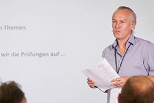Ein Mann steht vor einer Leinwand und redet. Er hat mittellange graue Haare und trägt ein Hemd. Vor ihm sind Zuhörer zu sehen.