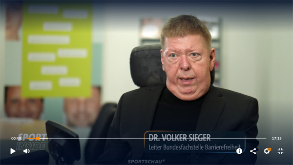 Ein Mann mit kurzen blonden Haaren spricht. Er trägt ein schwarzes Sakko und einen schwarzen Rollkragenpullover.