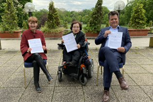 Zwei Frauen und ein Mann sitzen draußen auf einer Terrasse. Alle drei halten ein DIN A 4 Blatt in der Hand. Die Frau in der Mitte sitzt im Rollstuhl.