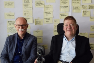 Peter Grothues, Direktor DRV-KBS, links, und Dr. Volker Sieger, Leiter der Bundesfachstelle Barrierefreiheit, rechts, sitzen vor einer Metaplanwand mit beschrifteten Karteikarten und schauen in die Kamera. Foto: Bundesfachstelle Barrierefreiheit/Miesner
