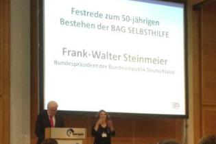 Bundespräsident Frank-Walter Steinmeier steht auf einer Bühne, neben ihm eine Gebärdensprachdolmetscherin. Im Hintergrund steht: Festrede zum 50-jährigen Bestehen der BAG SELBSTHILFE. Frank-Walter Steinmeier Foto: Bundesfachstelle Barrierefreiheit/Miesner