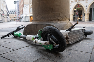 Das Foto zeigt einen liegenden E-Roller, der an einer Säule liegt. Der Gehweg ist Teil einer Fußgängerzone, im Hintergrund sind Geschäfte zu sehen. Foto: DBSV/Ziebe