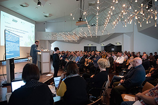 Das Publikum im Saal, Herr Wilhelm redet auf der Bühne - Foto: Bundesfachstelle Barrierefreiheit/Angelika Stehle