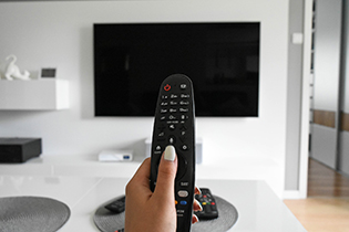 Eine Hand hält eine Fernbedienung, dahinter ist ein Fernseher zu sehen.