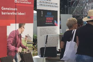 Das Bild zeigt den Informationsbereich der Bundesfachstelle Barrierefreiheit auf einem Stand des BMAS. Im Hintergrund befinden sich zwei Banner mit Schriftzügen. Rechts im Bild sind zwei Frauen von hinten zu sehen.